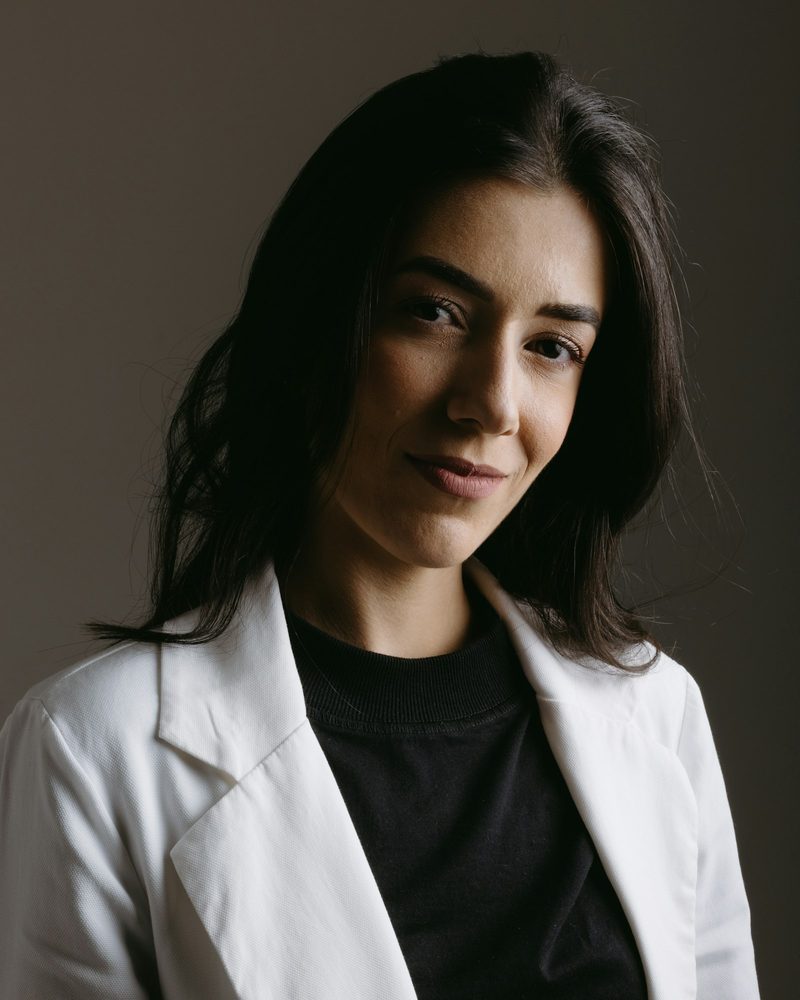 Portrait von Dr. Laura Sander mit langen dunklen Haaren, weißem Kittel und schwarzem Shirt vor dunklem Hintergrund