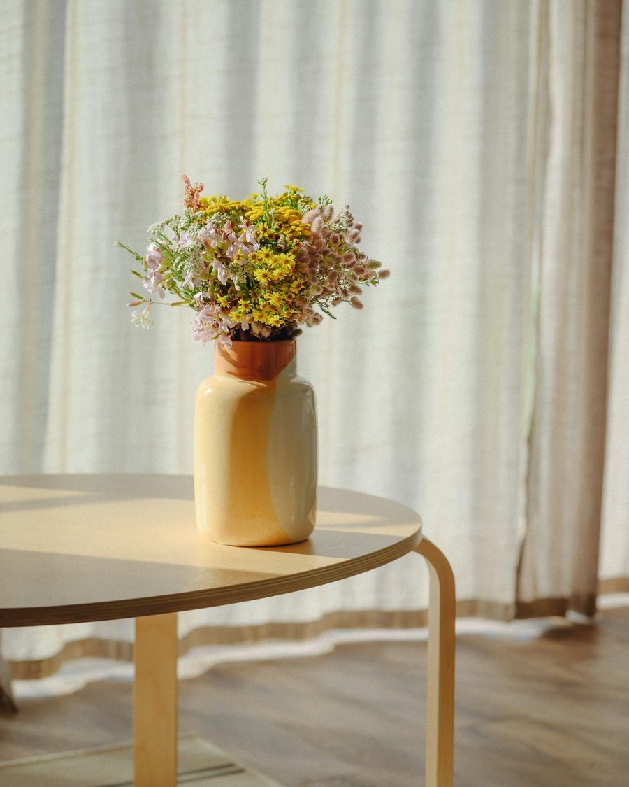 Heller Raum mit hellen Vorhängen, rundem Holztisch und einem Strauß Wiesenblumen in einer Vase