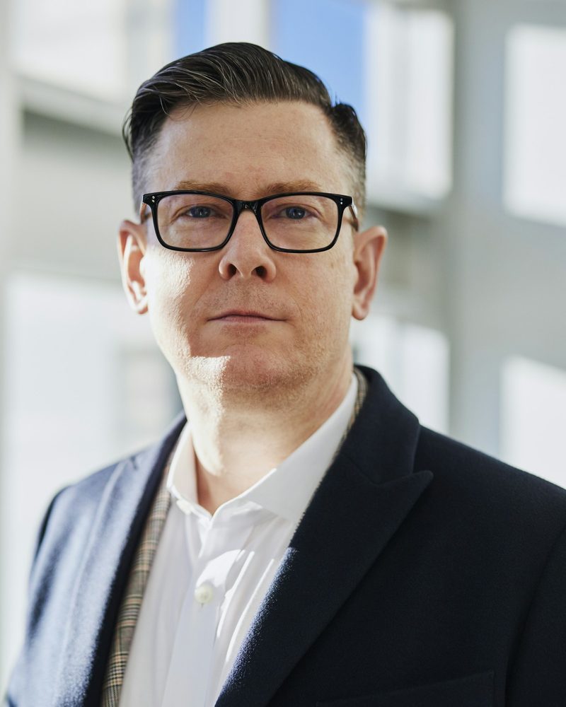 Portrait von Florian Amann mit Brille, dunklem Sakko und weißem Hemd vor einem hellen Fenster