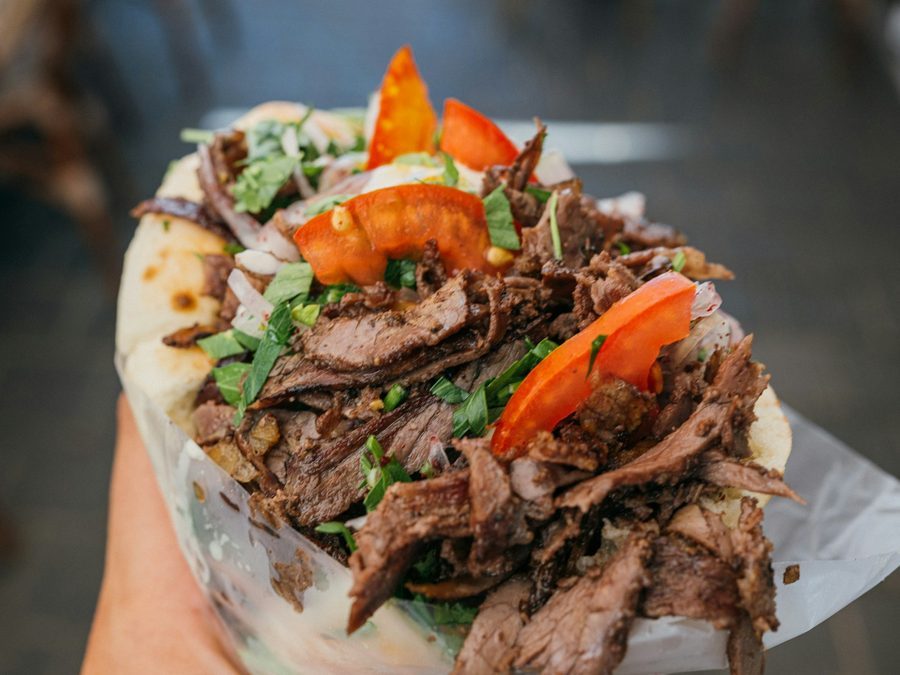 Hand hält einen Döner mit dünn geschnittenem Fleisch, Tomaten und Petersilie, im Hintergrund unscharf Tische und Stühle