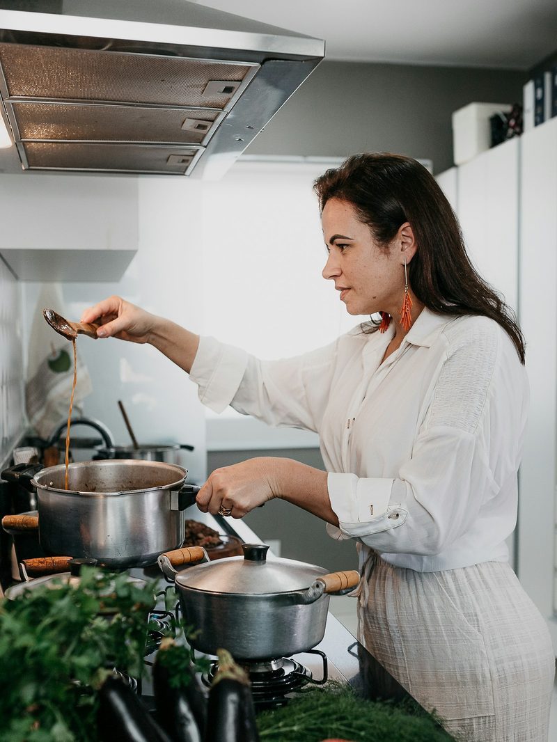 Portrait von Ayşe in weißer Bluse am Herd, sie rührt in einem Topf, davor Auberginen, Paprika und Kräuter