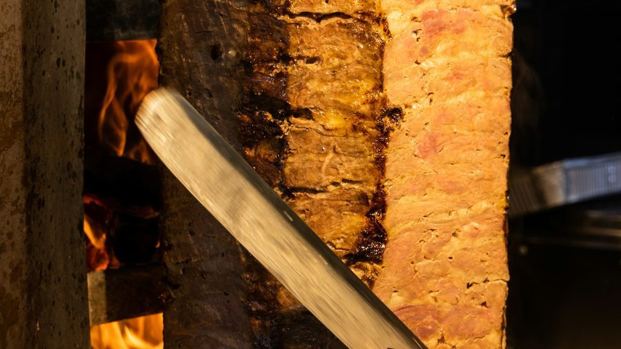 Dönerspieß am Grill, ein langes Messer schneidet dünne Scheiben ab, daneben die Flammen