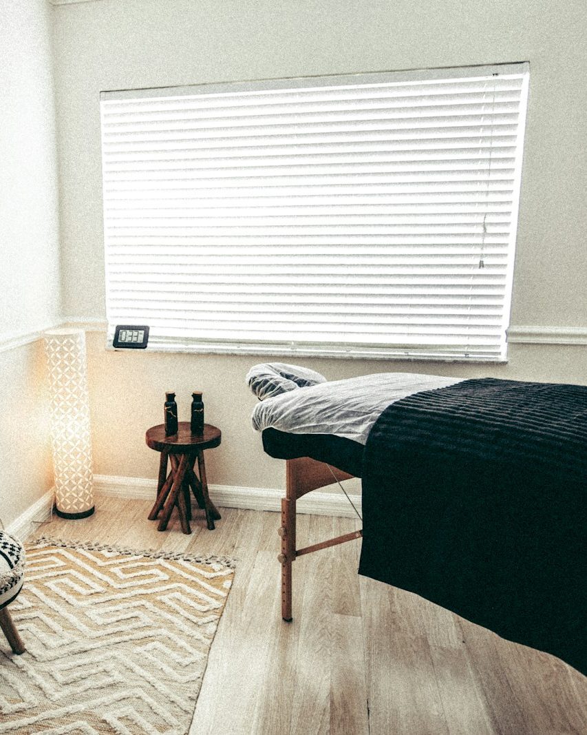 Heller Behandlungsraum im Haarentfernungs-Studio mit Liege unter dunkler Decke, Fenster mit weißen Jalousien, kleinem Holzhocker und Stehlampe