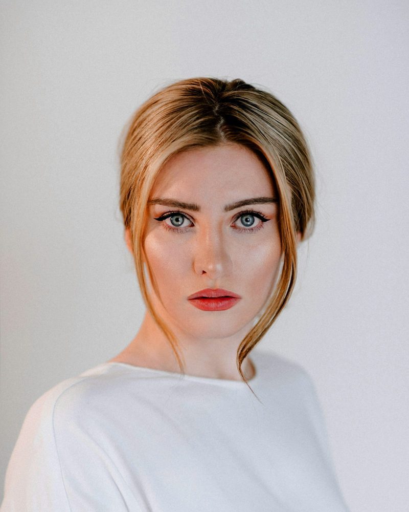 Portrait von Jana mit hochgesteckten blonden Haaren und weißem Oberteil vor hellgrauer Wand