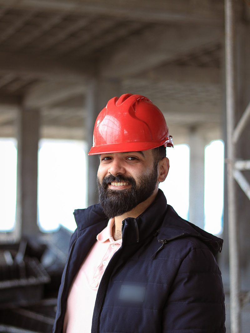 Portrait von Kevin, Geselle, mit Bart, rotem Schutzhelm und dunkler Arbeitsjacke auf einer Baustelle