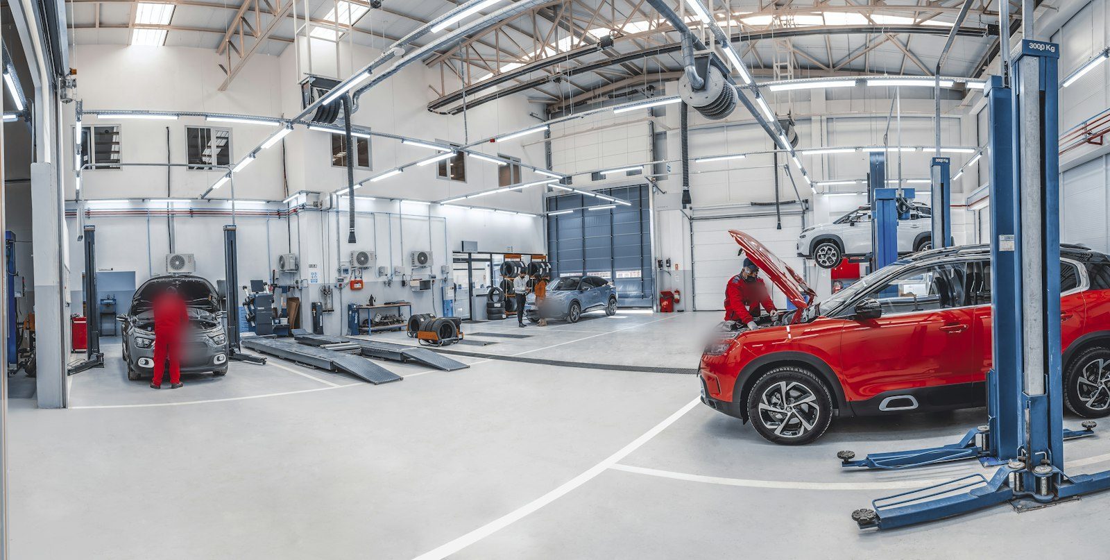 Moderne Website für eine Kfz-Werkstatt, Beispielansicht: Helle Werkstatthalle mit blauen Hebebühnen, ein roter Wagen mit offener Motorhaube, zwei Mechaniker in roter Arbeitskleidung