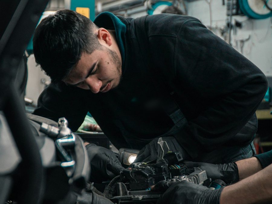 Junger Mechaniker in dunklem Pullover beugt sich mit Handschuhen über einen offenen Motorraum
