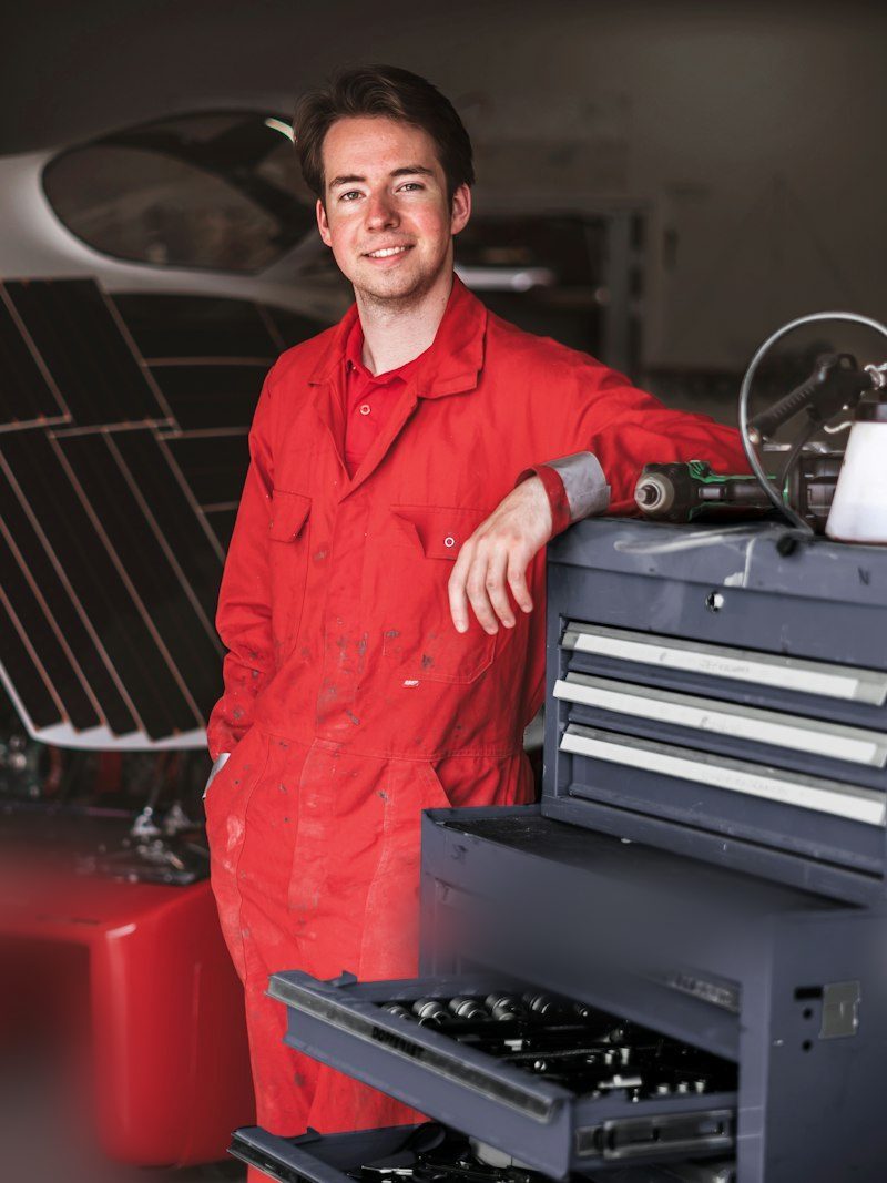 Portrait von Selim, Mechatroniker, in rotem Overall an einem Werkzeugwagen