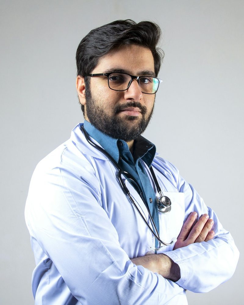 Portrait von Dr. med. Thomas Keller mit Brille, Bart, weißem Kittel und Stethoskop, Arme verschränkt, vor grauem Hintergrund