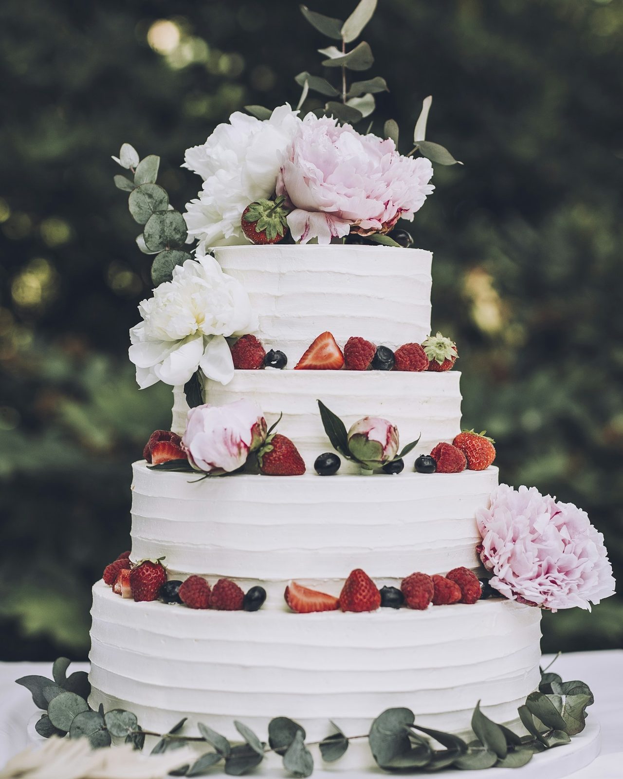 Moderne Website für eine Konditorei, Beispielansicht: weiße Hochzeitstorte mit vier Etagen, Himbeeren, Erdbeeren, Heidelbeeren und rosa Pfingstrosen vor grünem Garten