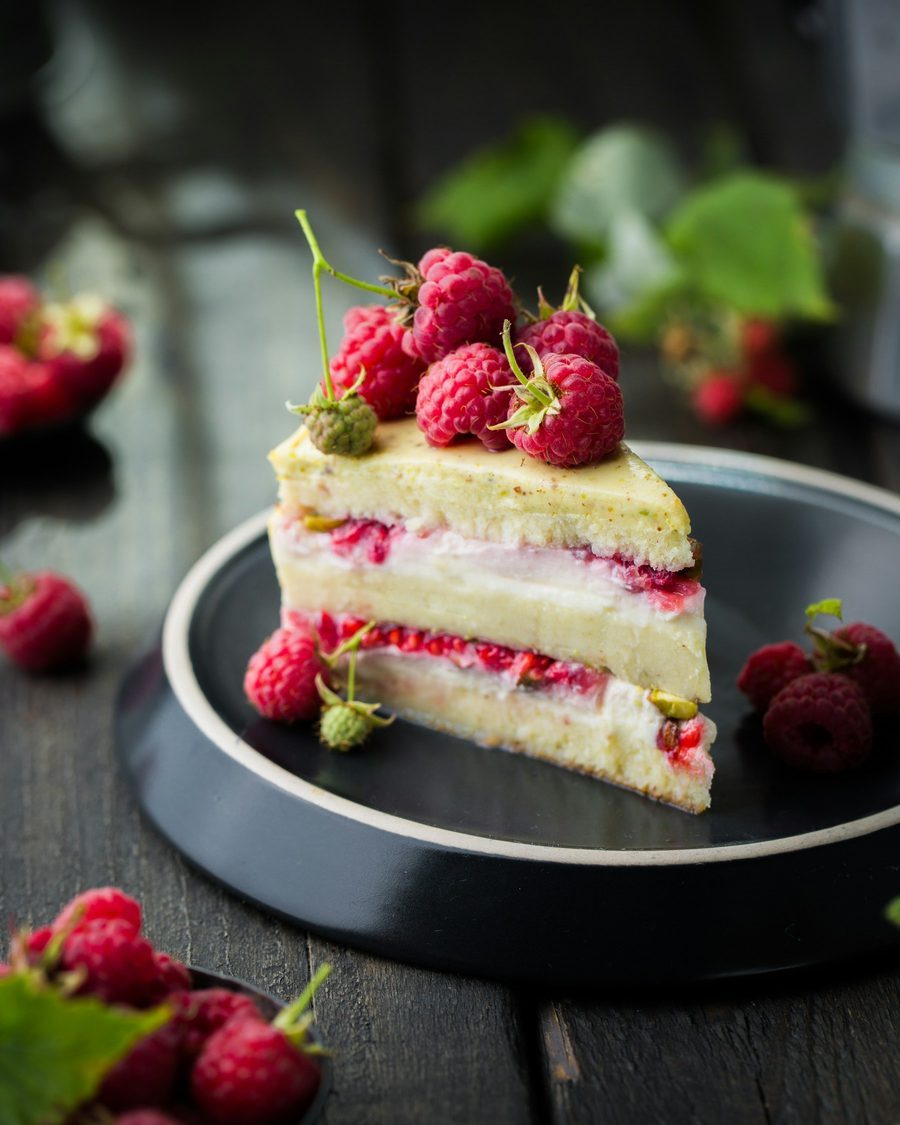 Stück Himbeertorte mit hellem Biskuit, Sahnecreme und frischen Himbeeren auf einem dunklen Teller