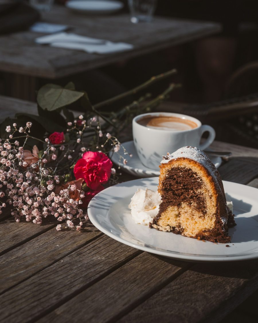 Holztisch im Café mit einer Tasse Cappuccino, einem Stück Marmorkuchen mit Sahne und einer roten Nelke