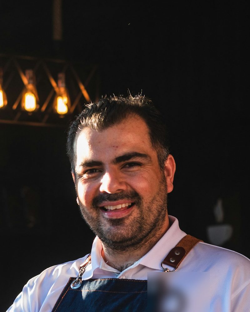 Portrait von Leo mit Bart, weißem Hemd und dunkler Schürze, lachend im Café