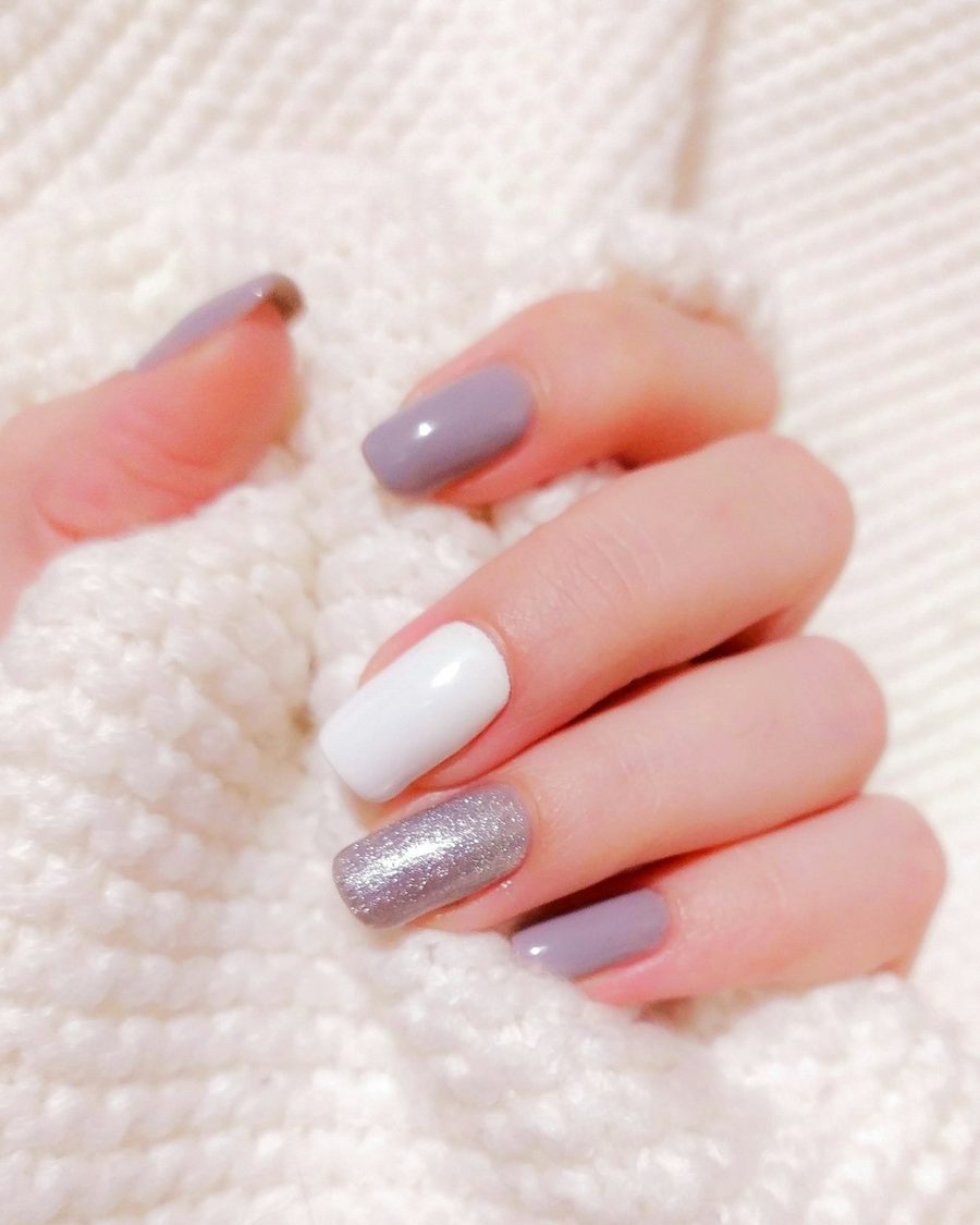 Hand mit Gelnägeln aus dem Nagelstudio in Flieder, Weiß und Silberglitzer auf einem weißen Strickpullover
