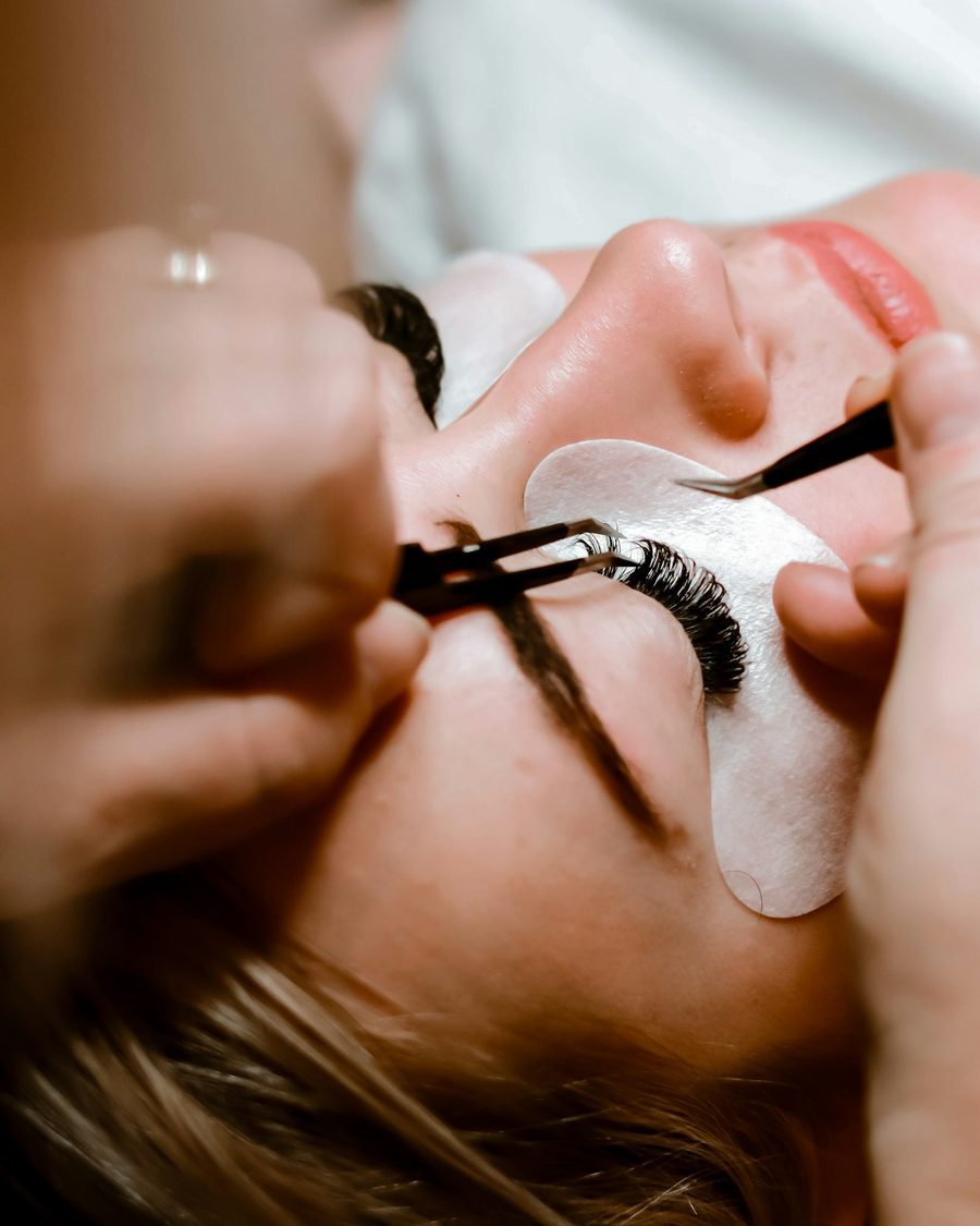 Wimpernstylistin setzt mit zwei Pinzetten einzelne Wimpern bei einer liegenden Kundin mit Augenpad