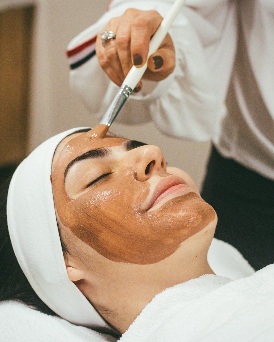 Kosmetikerin verteilt mit dem Pinsel eine Tonerde-Maske auf dem Gesicht einer Kundin mit weißem Haarband