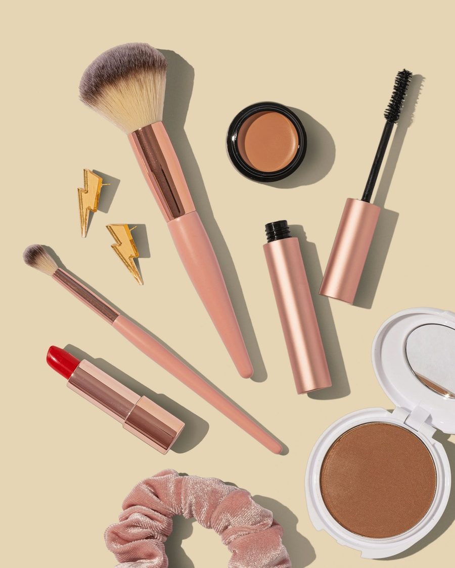 Pinsel, Mascara, Lippenstift und Puder in Rosé auf sandfarbenem Grund im Kosmetikstudio