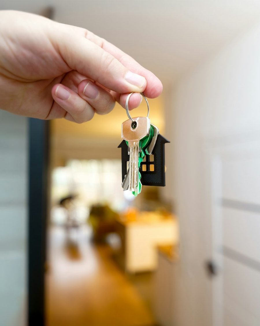 Hand hält einen Schlüsselbund mit hausförmigem Anhänger in einem hellen Flur, dahinter unscharf eine Wohnung