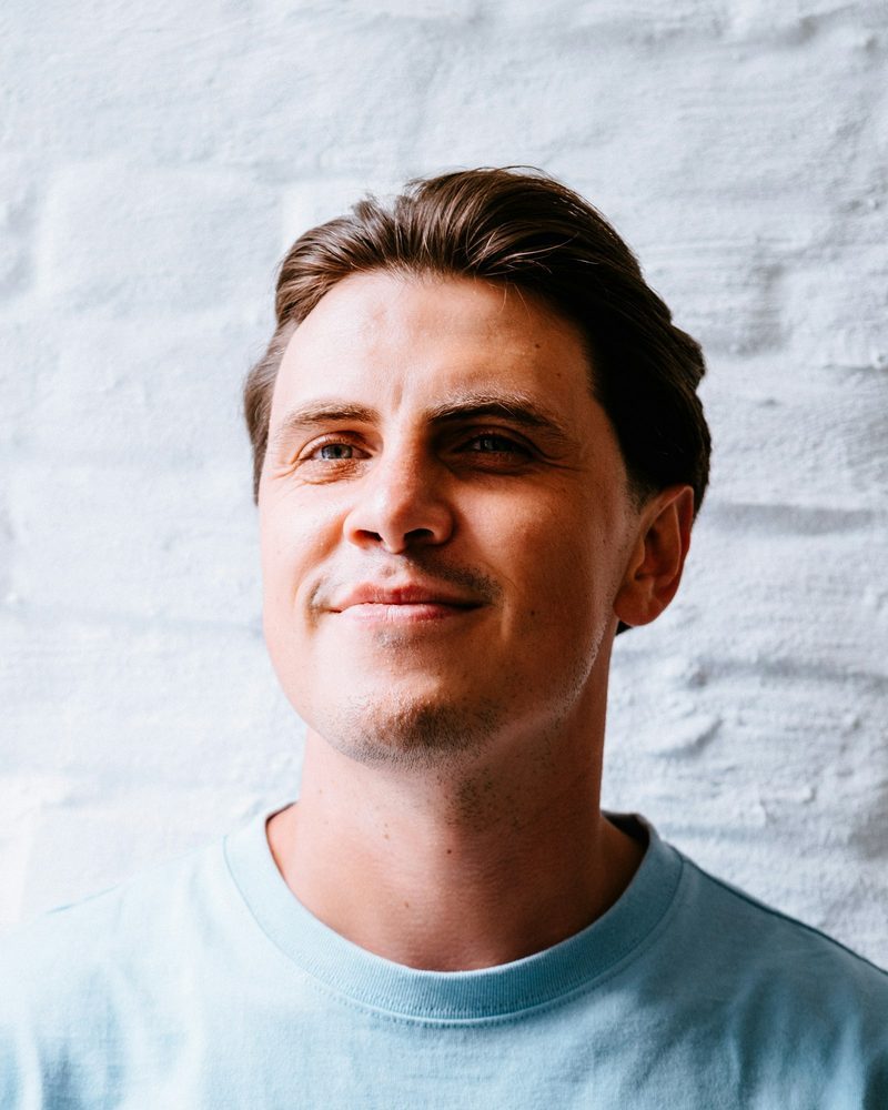 Portrait von Tom mit kurzen braunen Haaren und hellblauem T-Shirt vor einer weißen Ziegelwand