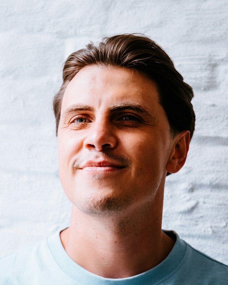 Portrait von Lukas, Sportphysiotherapeut, im hellblauen T-Shirt vor einer weißen Ziegelwand