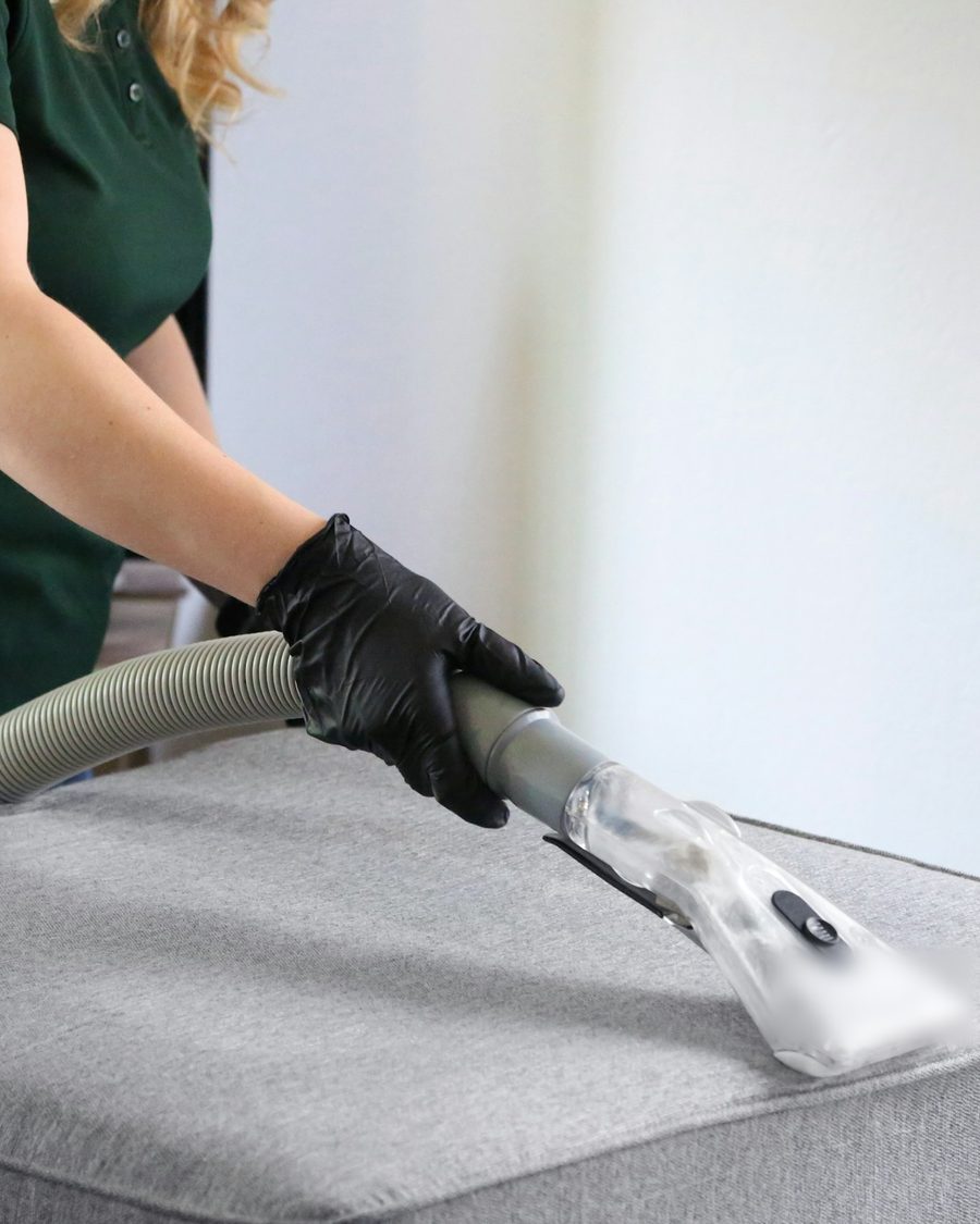 Mitarbeiterin im dunkelgrünen Poloshirt mit schwarzen Handschuhen reinigt ein graues Polster mit einer Absaugdüse