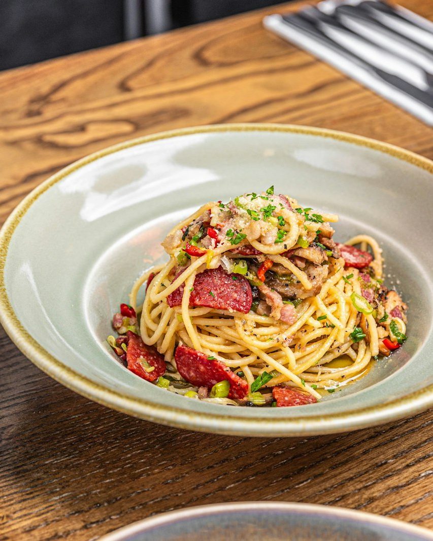 Teller Spaghetti mit Salami, Kräutern und Chili auf einem Holztisch, daneben ein Glas Spritz