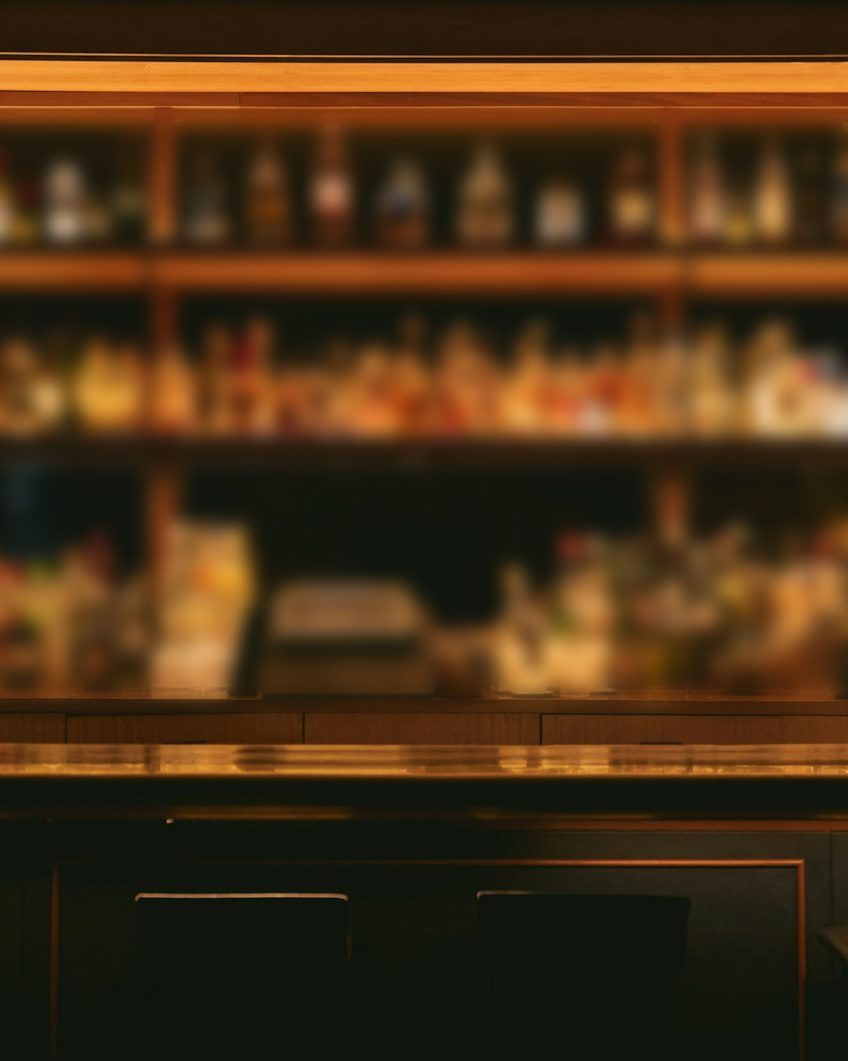 Bar in warmem Licht mit beleuchtetem Regal voller Flaschen, dunkle Barhocker am Tresen