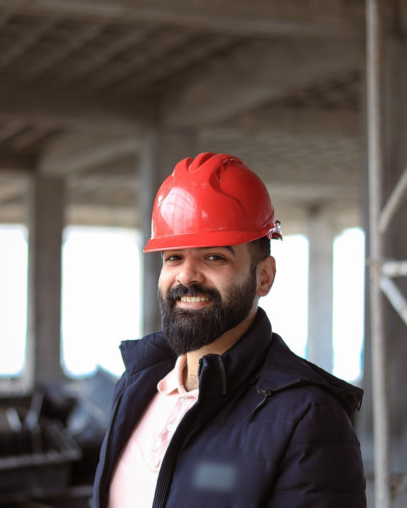 Portrait von Robert Kainz mit rotem Bauhelm, Vollbart und dunkler Jacke auf einer Baustelle