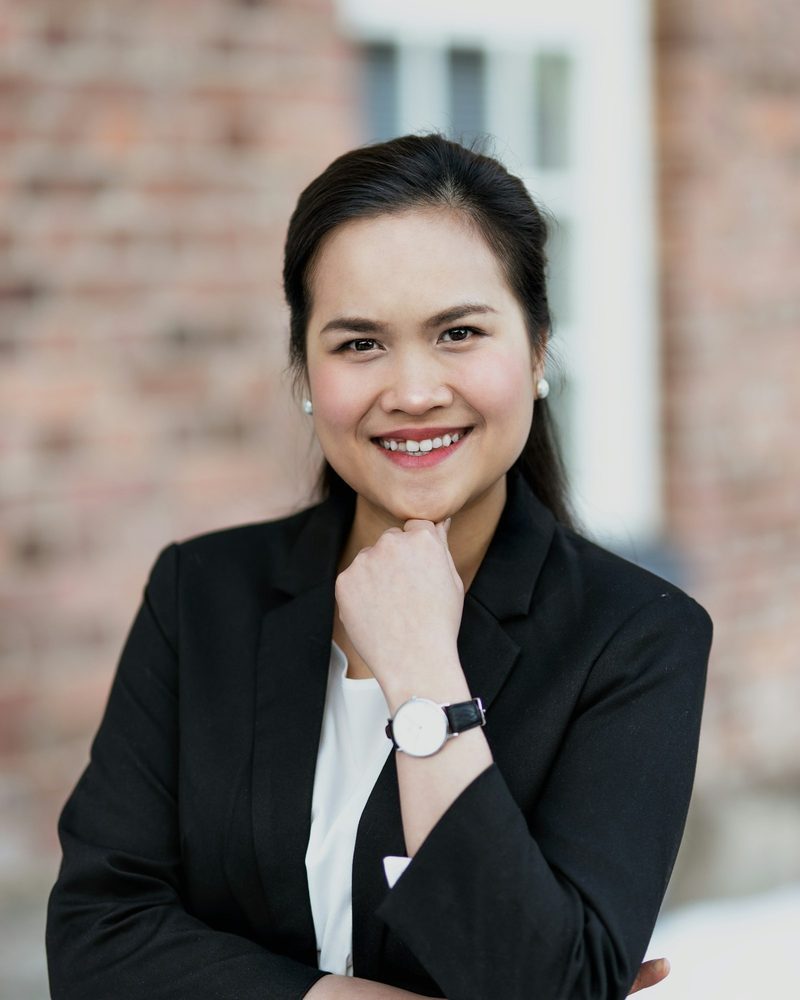 Portrait von Sandra mit dunklem Haar, schwarzem Blazer und Armbanduhr vor einer Ziegelwand