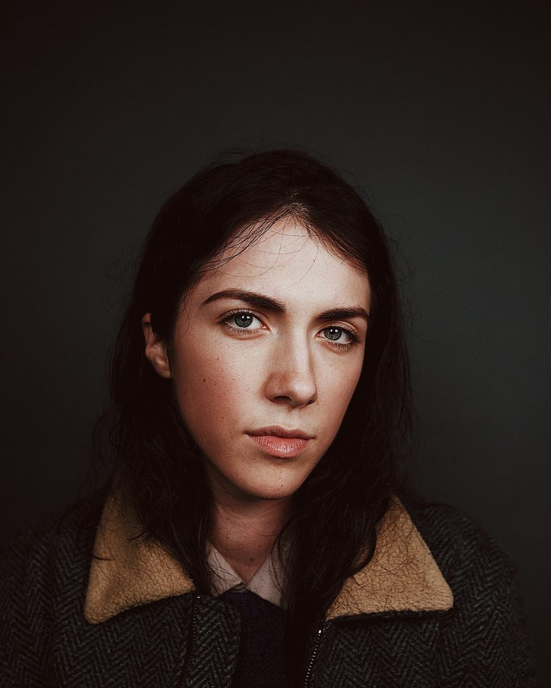 Portrait von Lisa mit dunklen langen Haaren und Wolljacke mit hellem Kragen vor dunklem Hintergrund