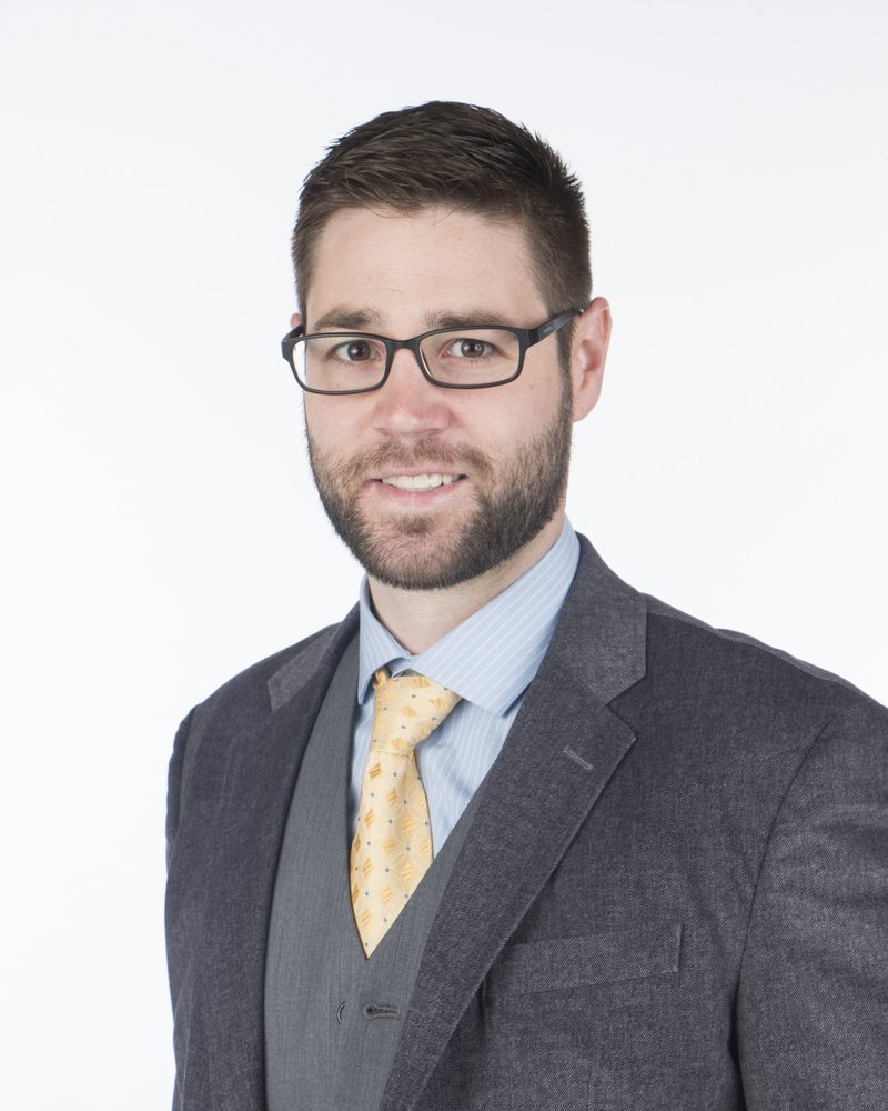 Portrait von Daniel mit Brille, Bart, grauem Anzug und gelber Krawatte vor hellem Hintergrund