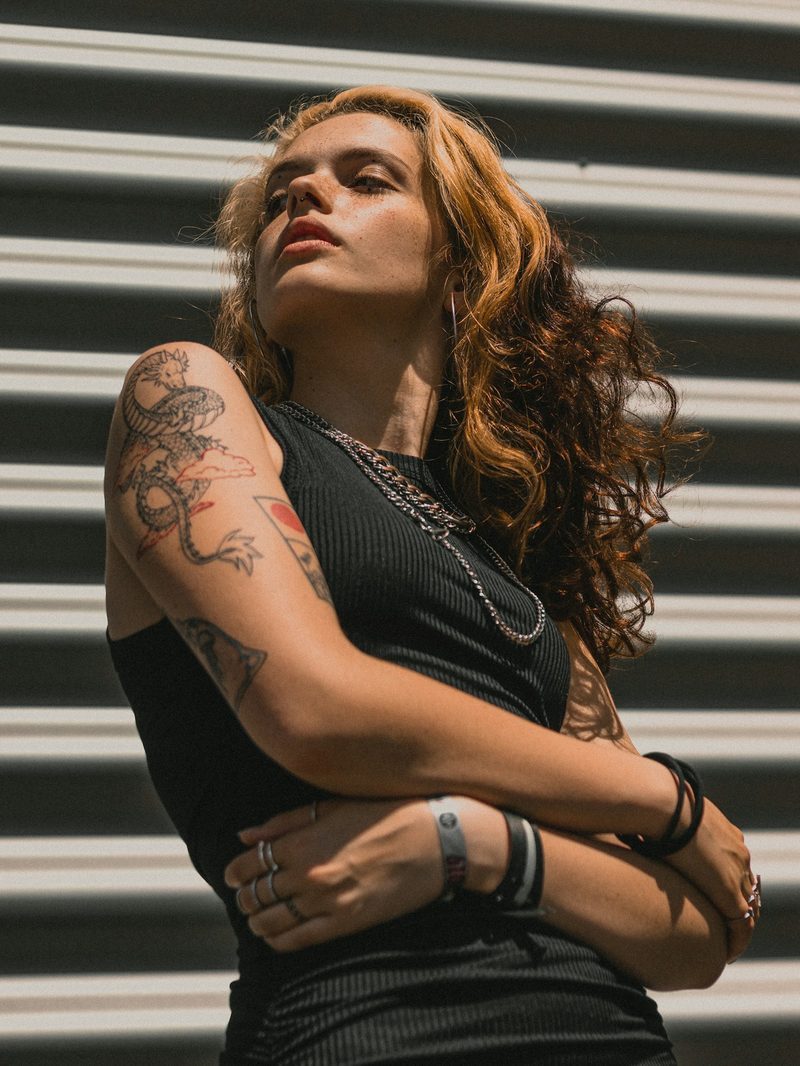 Portrait von Mira mit langen lockigen Haaren und tätowierten Armen vor einer hellen Metallwand