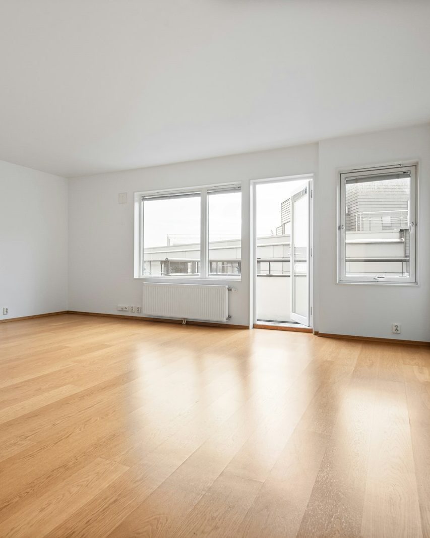 Leeres, helles Wohnzimmer mit Holzparkett, weißen Wänden, Heizkörper und Balkontür
