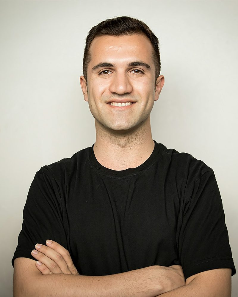 Portrait von Jonas, Teamleiter, im schwarzen T-Shirt mit verschränkten Armen vor einer hellen Wand