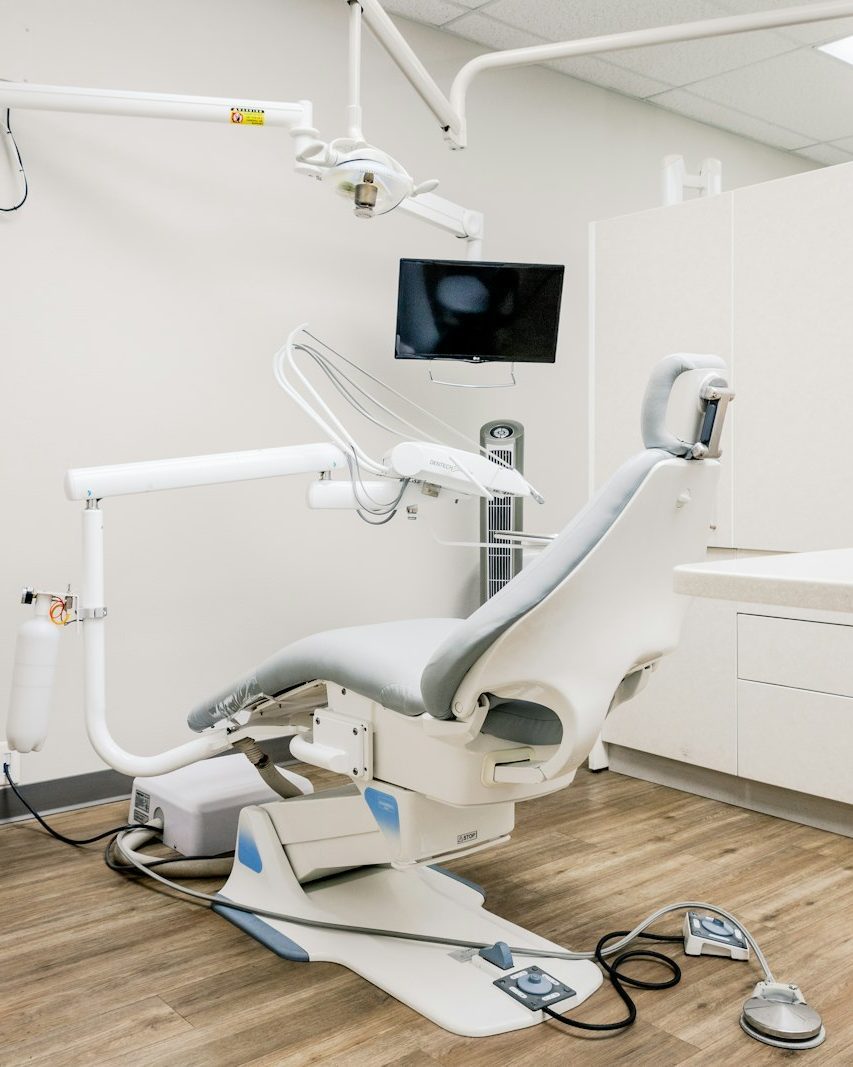 Helles Behandlungszimmer einer Zahnarztpraxis mit grauem Behandlungsstuhl, Lampe, Bildschirm und Holzboden